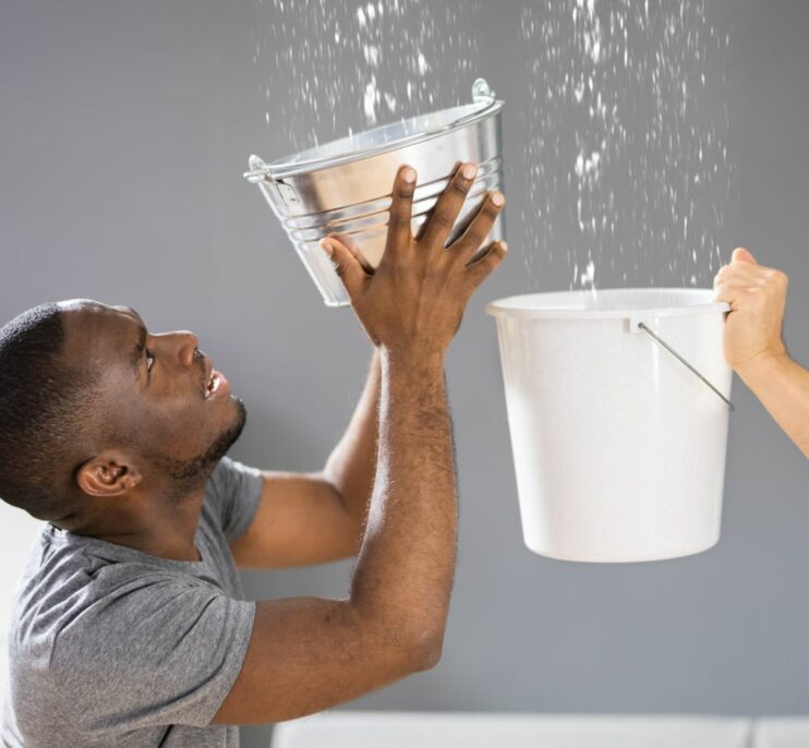 Two men attempting to catch water leaking from the ceiling with buckets while one makes an urgent phone call. Signs of a serious roof leak that requires immediate repair. Empire Contractors offers professional roof leak detection and emergency repair services to protect homes from water damage.
