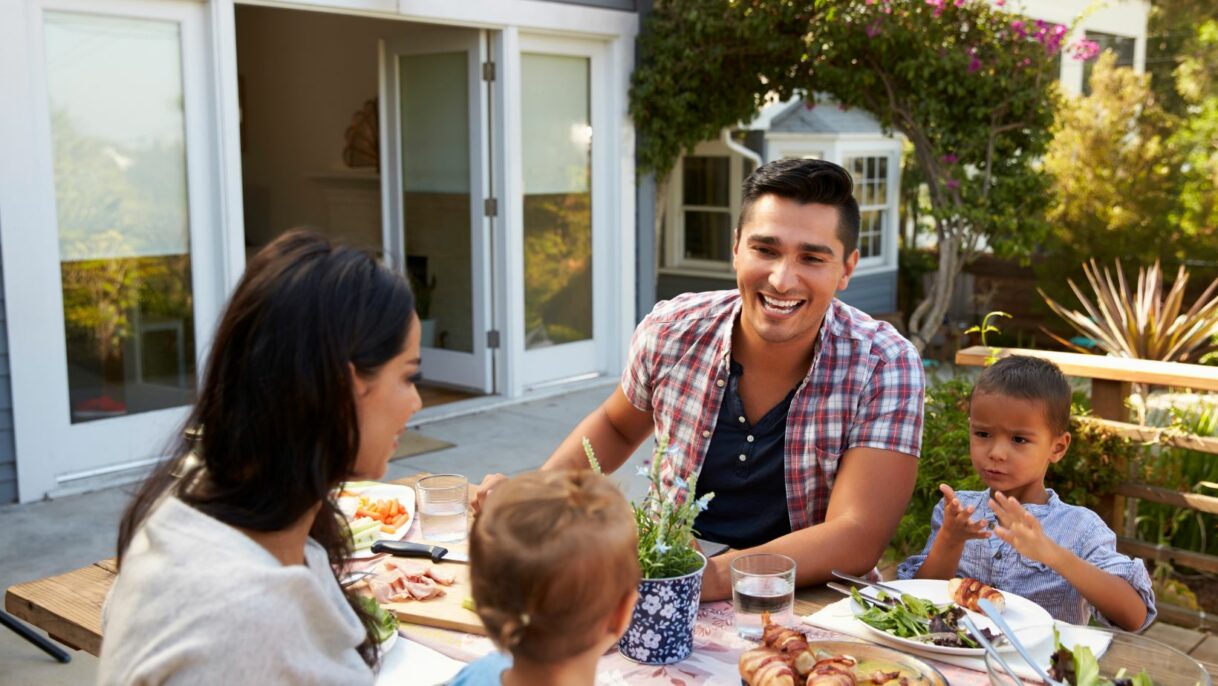 Happy family enjoying an outdoor meal on a well-built patio with sliding glass doors leading to a modern home. Beautiful backyard renovation with landscaping and wooden deck. Empire Contractors specializes in high-quality patio and deck construction.