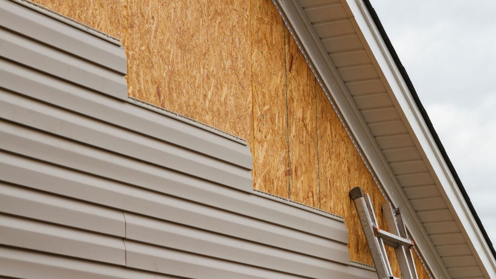 Partially installed beige vinyl siding on a home exterior, revealing the wooden sheathing underneath. A ladder is positioned for ongoing construction or repair. Empire Contractors provides professional siding installation and replacement for durable, weather-resistant home exteriors.