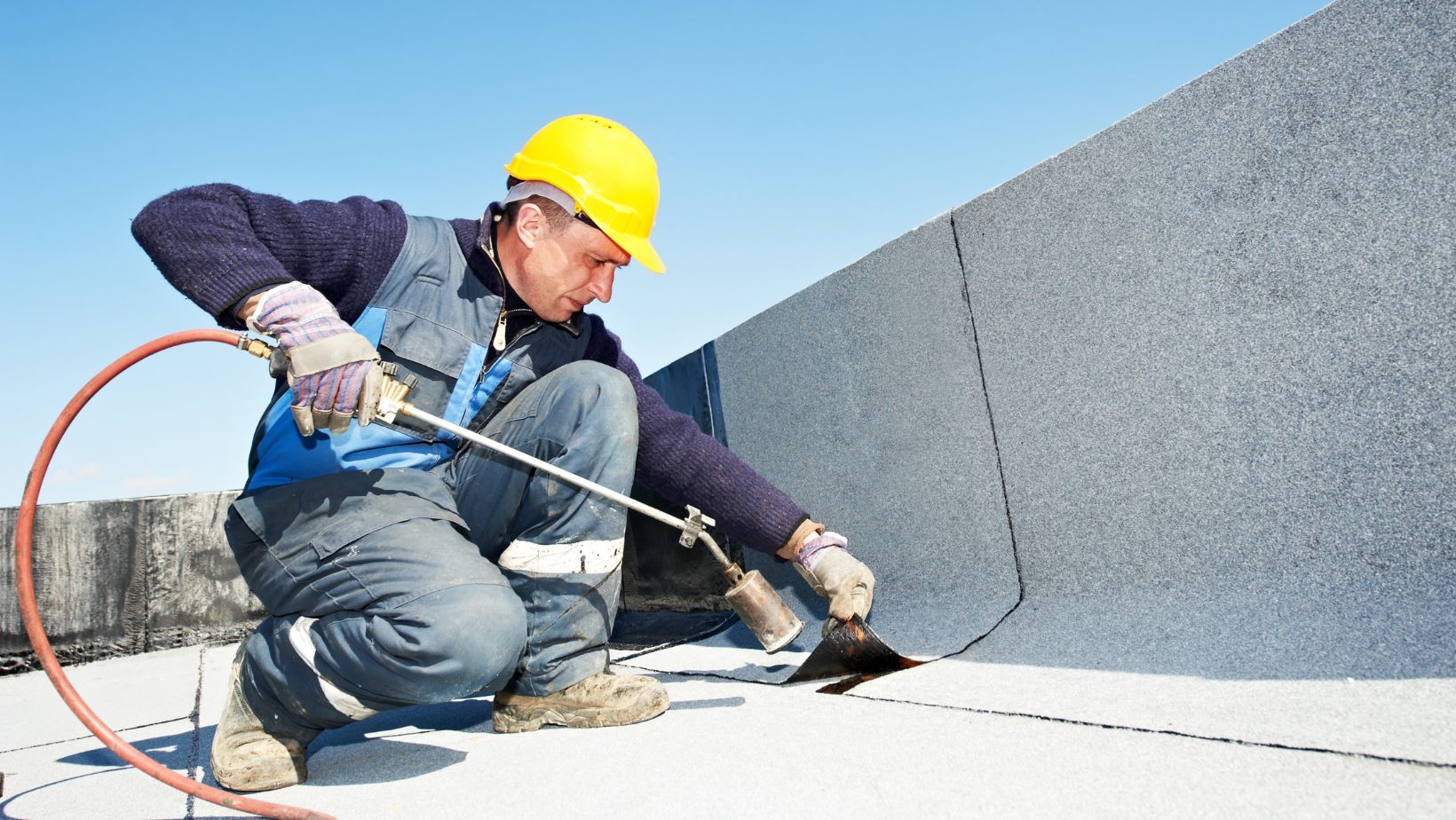 Roofing contractor wearing a yellow hard hat and protective gear applies a torch-down membrane to a flat commercial roof for waterproofing. Empire Contractors specializes in professional flat roof installation and repair for long-lasting durability and weather resistance.